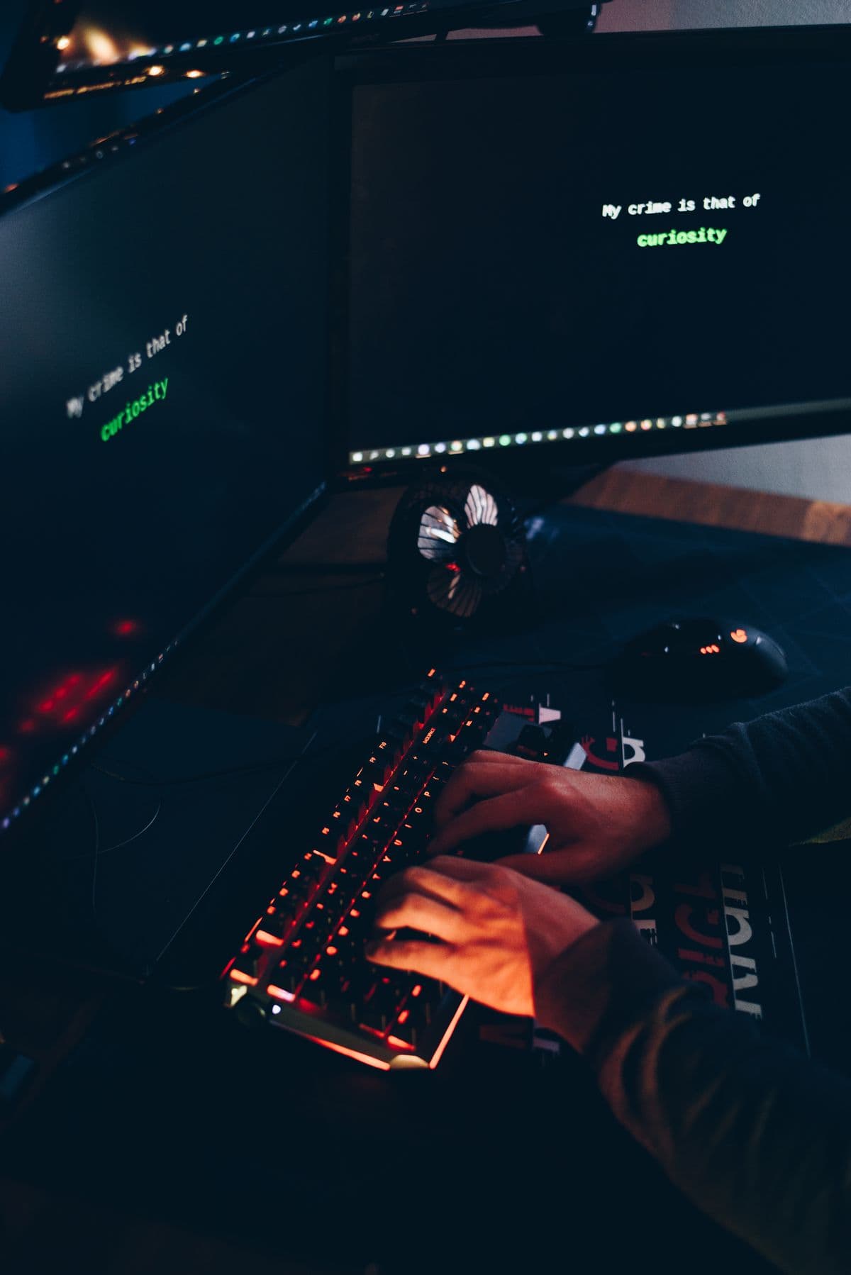 Hands typing on a backlit keyboard in a dark room lit by two monitors