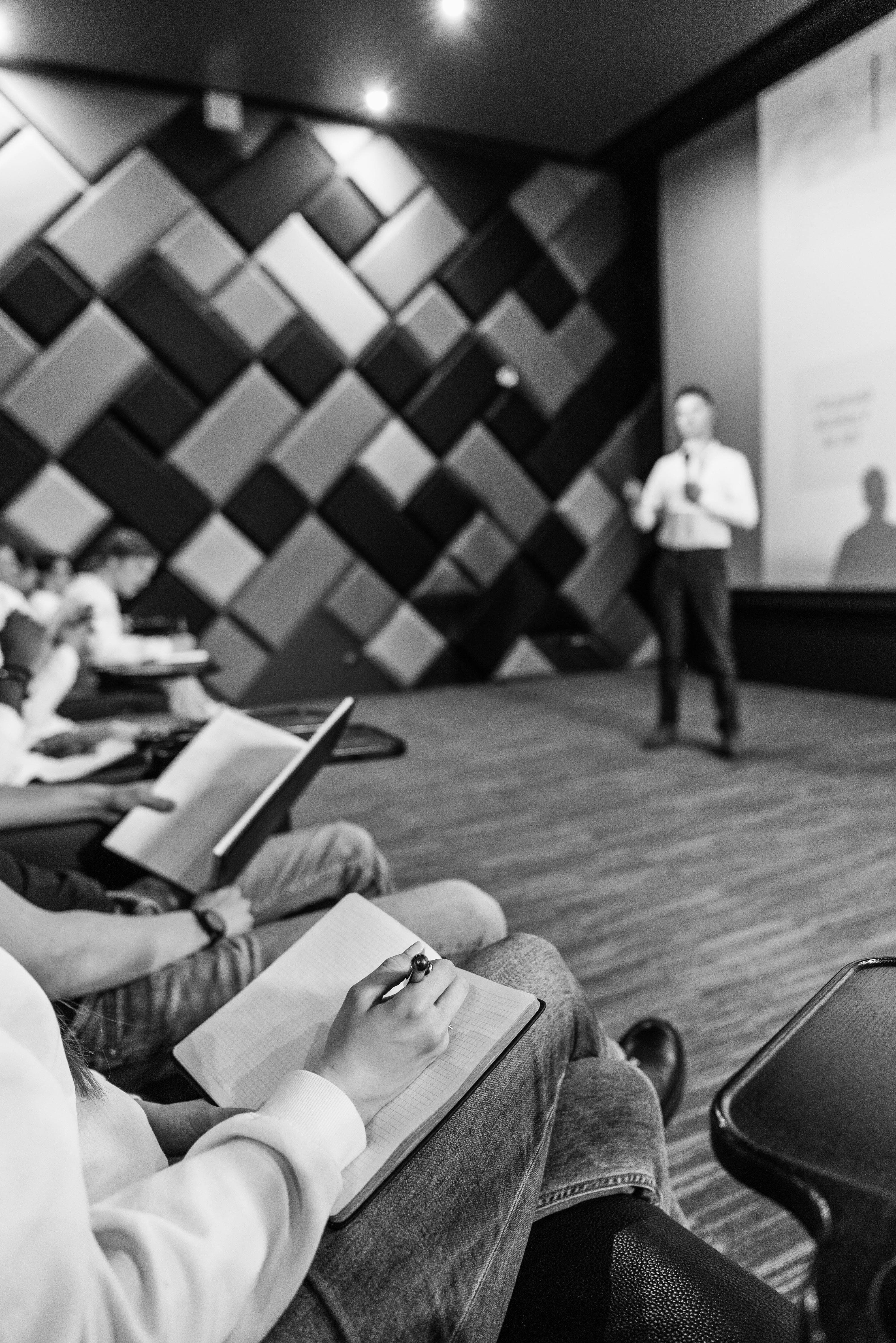 A speaker presenting at a startup meetup while attendees take notes, in black and white